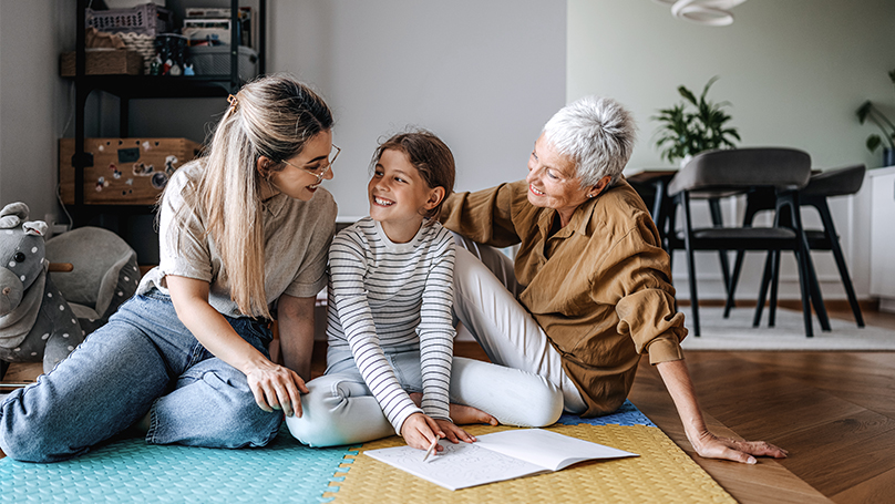Mother, grandmother, and granddaughter on a play mat, coloring in a coloring book.