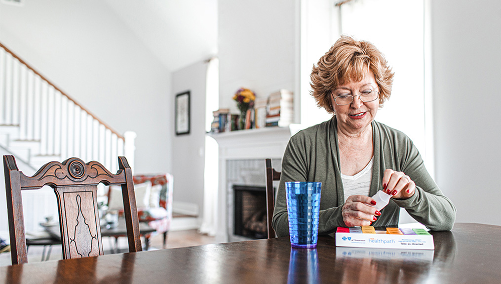 A senior woman refilling her pill planner at her dining room table.