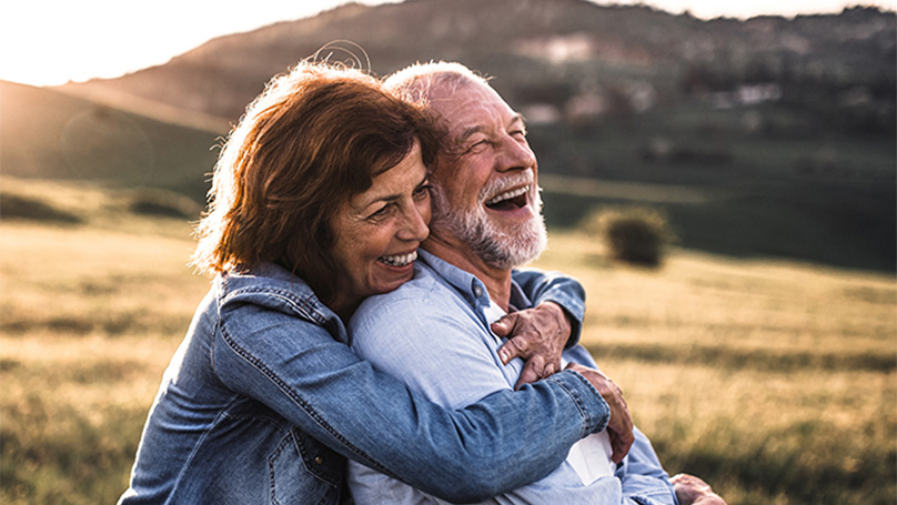 A senior woman hugs a senior man while on an outdoor walk.