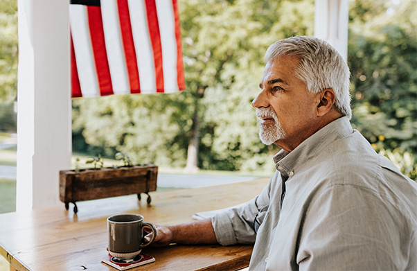 An older man sitting on his front porch with an American flag in the background.